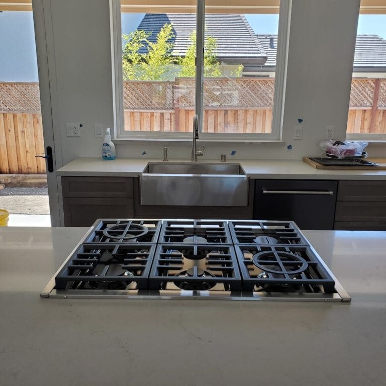 Picture taken from a large kitchen island. A 6 burner built-in counter gas range takes center stage with a stailess steel sink beyond it.